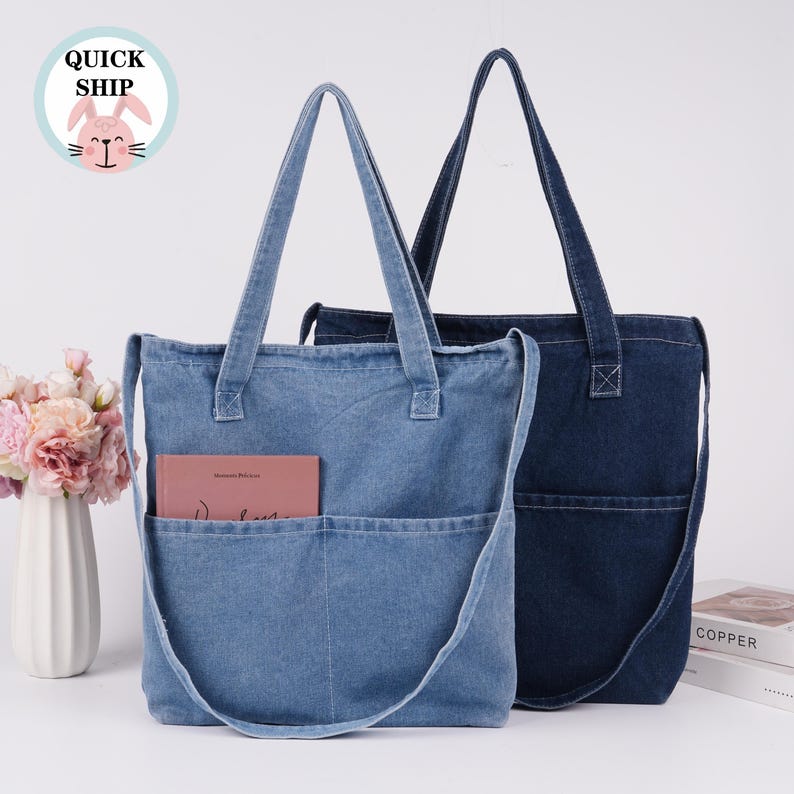 Peut inclure: Deux sacs fourre-tout en jean, un bleu clair et un bleu fonc&eacute;, avec de longues sangles et des poches avant. Le sac bleu clair contient un livre dans sa poche. Un autocollant "QUICK SHIP" avec un lapin est en haut &agrave; gauche.