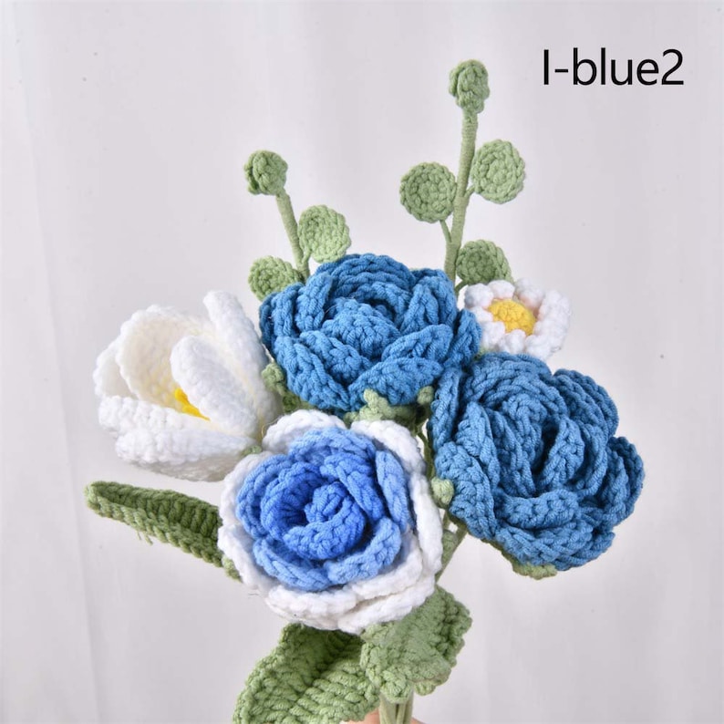 May include: A bouquet of crocheted flowers in shades of blue and white. The bouquet includes three blue roses, one white rose, and one white flower with a yellow center. The flowers are attached to green crocheted stems.