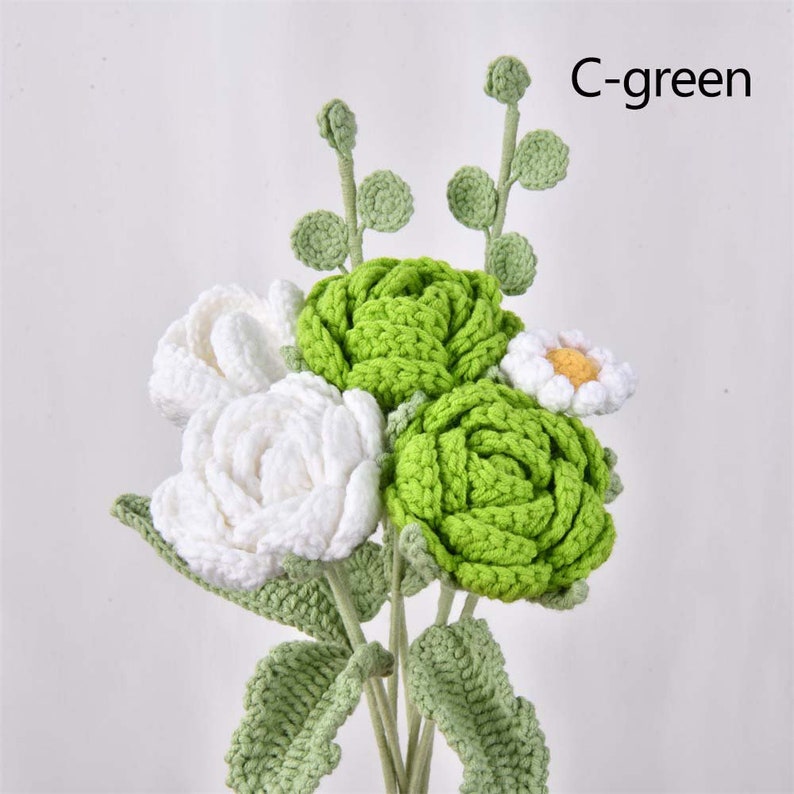 May include: A bouquet of crocheted flowers in white and green. The bouquet includes three white roses, two green roses, and a small white daisy. The flowers are arranged in a vase and have green leaves.