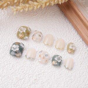 May include: A collection of ten artificial nails in various shapes and designs. Some nails feature floral patterns in white, gold, and green, while others are solid colors. The nails are arranged on a textured white surface.