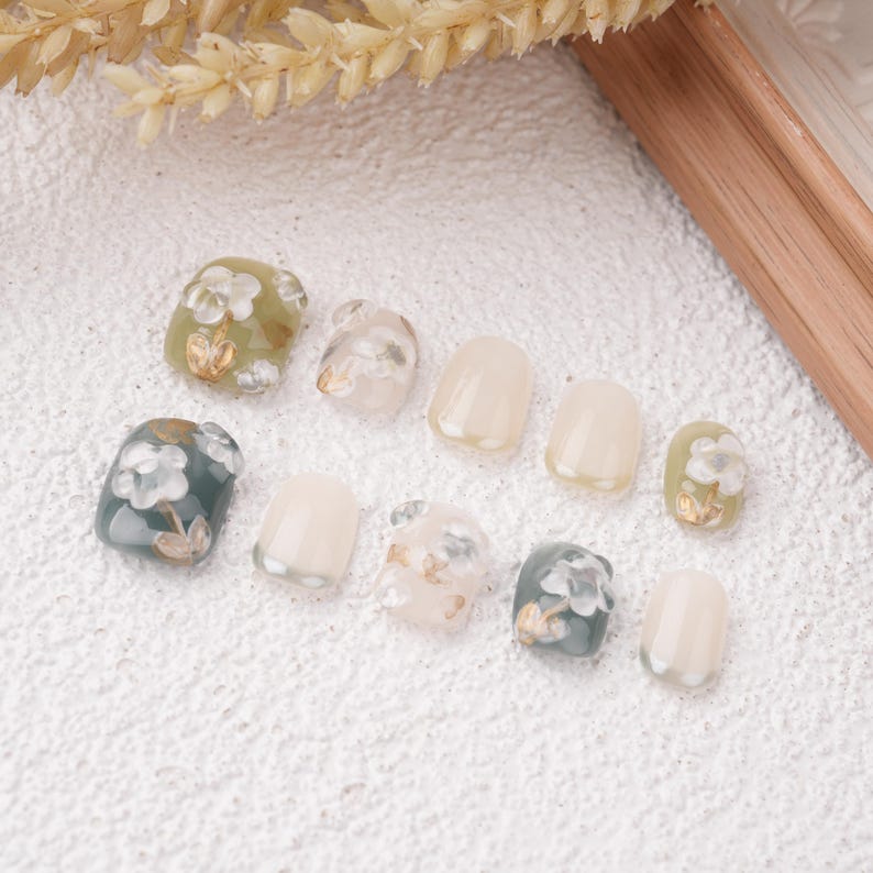 May include: A collection of ten artificial nails in various shapes and designs. Some nails feature floral patterns in white, gold, and green, while others are solid colors. The nails are arranged on a textured white surface.