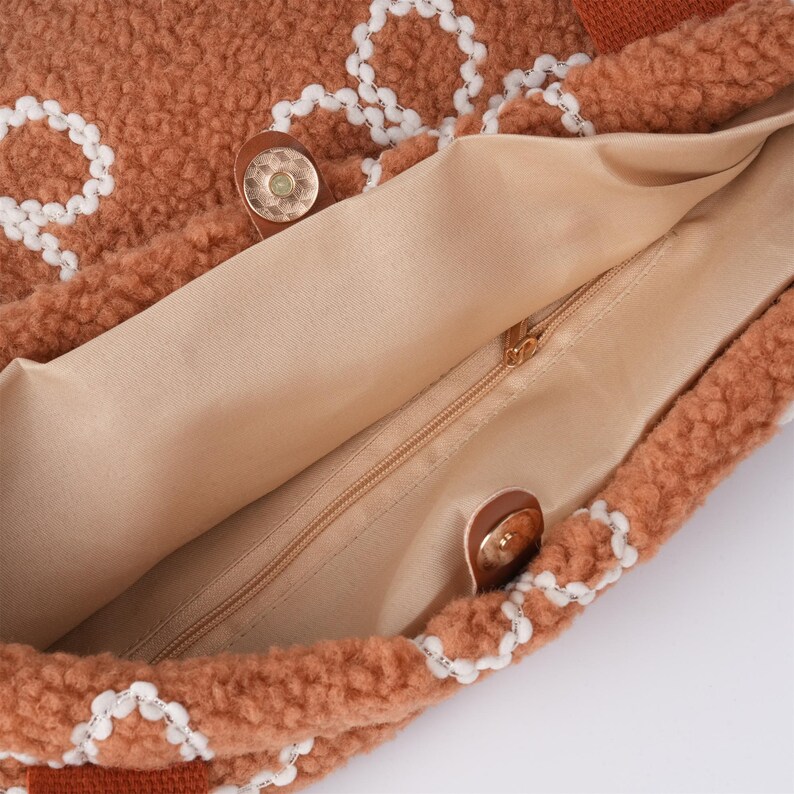 May include: The inside of a brown fluffy tote bag with a white embroidered design. The bag has a zip closure and a brown fabric lining.