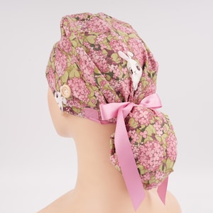 May include: A scrub cap featuring a floral pattern of pink hydrangea clusters and green leaves on a dark background, interspersed with white rabbit illustrations. The cap includes a light pink satin ribbon tie and a side button for mask attachment.