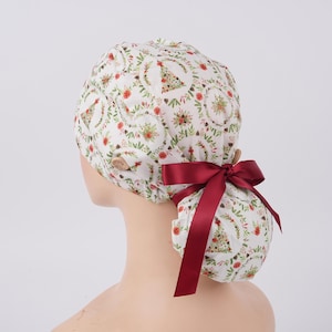 May include: A white scrub cap featuring a repeating pattern of green pine trees, red flowers, and brown pinecones. The cap includes two light-colored buttons on the sides and a dark red satin ribbon tied into a bow at the back to secure the fabric.