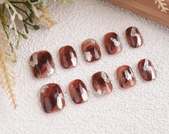 May include: A set of ten press-on nails. Each nail is a mix of brown and tan hues with silver foil accents. The nails have a glossy finish and a rounded shape, suitable for a manicure.