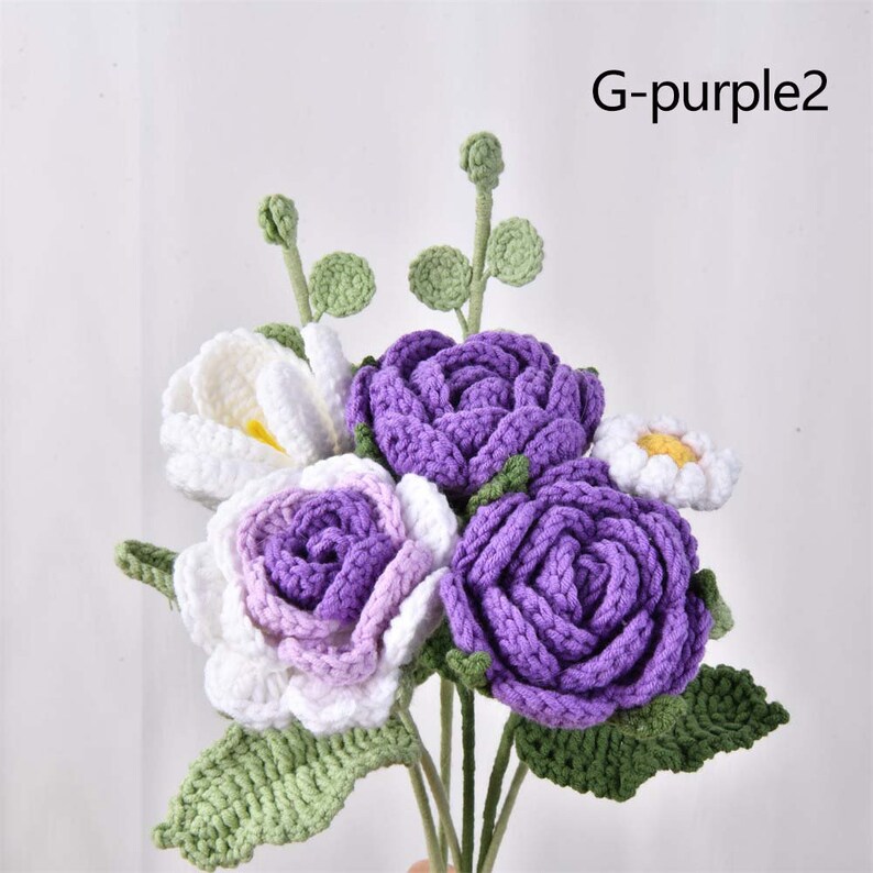 May include: A bouquet of crocheted flowers in shades of purple, white, and green. The bouquet includes three purple roses, one white rose with purple accents, and one white daisy. The flowers are arranged in a vase and have green leaves.