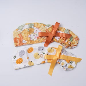 May include: Two scrub caps featuring autumn-themed patterns. The top cap displays orange pumpkins and white rabbits with a burnt orange ribbon. The bottom cap features a white background with small orange and gray pumpkins and a yellow ribbon.