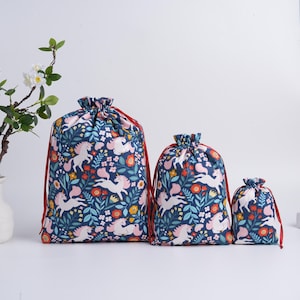 May include: Three drawstring bags of varying sizes featuring a dark blue fabric base with a repeating pattern of white unicorns, red and yellow flowers, and green leaves. Each bag is cinched at the top with thin red cord strings for closure.