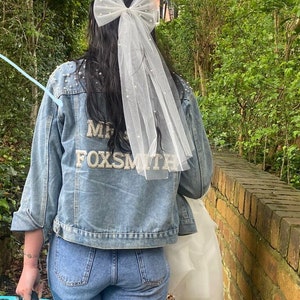 May include: A light blue denim jacket with pearl detailing and the name "MRS FOXSMITH" on the back. The person is wearing a white bow veil and a tiara. The person is carrying a turquoise suitcase and a white garment bag.