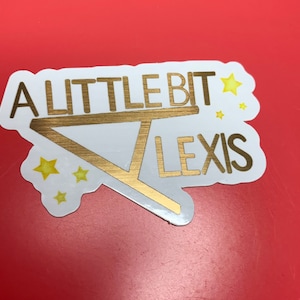 Op de afbeelding: Een witte sticker met gouden letters die "A Little Bit Alexis" lezen met een gouden driehoeksvorm en gele sterren.