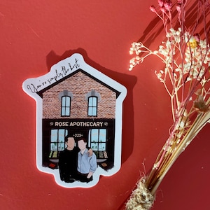 Puede incluir: Una pegatina de un edificio de ladrillo con una fachada de tienda que dice "ROSE APOTHECARY" y el texto "You're simply the best" sobre la fachada. Dos personas están de pie frente a la tienda.