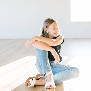 May include: A young person wearing a black t-shirt and blue jeans sits on a wooden floor, smiling and looking to the right. They are wearing white sandals with a thick platform.