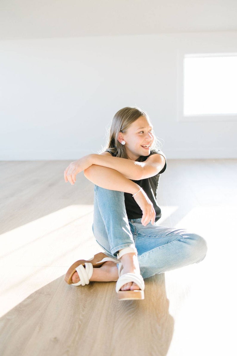May include: A young person wearing a black t-shirt and blue jeans sits on a wooden floor, smiling and looking to the right. They are wearing white sandals with a thick platform.