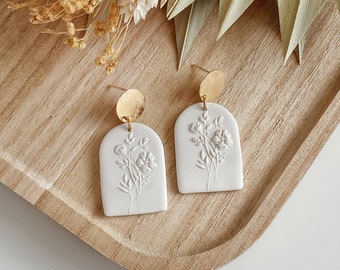 May include: A pair of white ceramic earrings with a floral design and gold accents. The earrings are shaped like arches and feature delicate floral patterns.