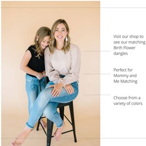 May include: Two people, a woman and a girl, are wearing jeans and smiling. The woman is wearing a light pink top and the girl is wearing a black top. They are both wearing earrings. The text in the image reads: "Visit our shop to see our matching Birth Flower dangles. Perfect for Mommy and Me Matching. Choose from a variety of colors."