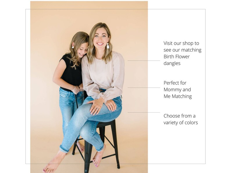 May include: Two people, a woman and a girl, are wearing jeans and smiling. The woman is wearing a light pink top and the girl is wearing a black top. They are both wearing earrings. The text in the image reads: "Visit our shop to see our matching Birth Flower dangles. Perfect for Mommy and Me Matching. Choose from a variety of colors."