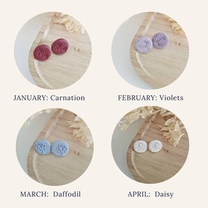 May include: Four pairs of small, round, stud earrings with floral designs. The earrings are displayed on a wooden surface. The colors of the earrings are burgundy, lavender, light blue, and white. The text below each pair of earrings reads "JANUARY: Carnation", "FEBRUARY: Violets", "MARCH: Daffodil", and "APRIL: Daisy".