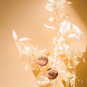 May include: Two brown clay earrings with a delicate floral design, displayed on a white dried flower branch against a light beige background.