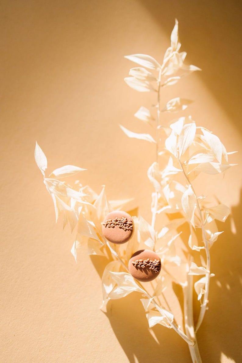 May include: Two brown clay earrings with a delicate floral design, displayed on a white dried flower branch against a light beige background.