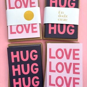 Può includere: Quattro pile di biglietti d'auguri con sfondi rosa e neri. Le carte presentano le parole "LOVE" e "HUG" in grandi lettere rosa. Una pila ha il testo "if in doubt create." su una fascia bianca.