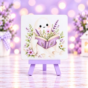 May include: A square white sign featuring a cartoon ghost holding an open purple book, surrounded by lavender and pink flowers, displayed on a small light purple wooden easel stand.