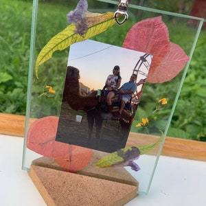 Puede incluir: Un marco de fotos de vidrio con un diseño de flores prensadas. El marco está decorado con hojas y flores secas en tonos de rosa, amarillo y morado. Una foto de dos personas montando un camello está dentro del marco.