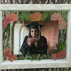 Puede incluir: Una fotografía enmarcada de una persona con un gorro y una bata de graduación, soplando confeti. El marco está decorado con flores y hojas prensadas.