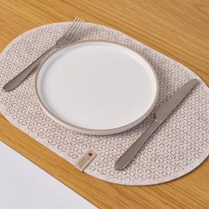 Puede incluir: Un mantel individual de color beige con un diseño geométrico, un plato blanco, un tenedor y un cuchillo. El mantel individual tiene una etiqueta que dice "EVADAY". El plato tiene un borde marrón claro. Los artículos están colocados sobre una mesa de madera.