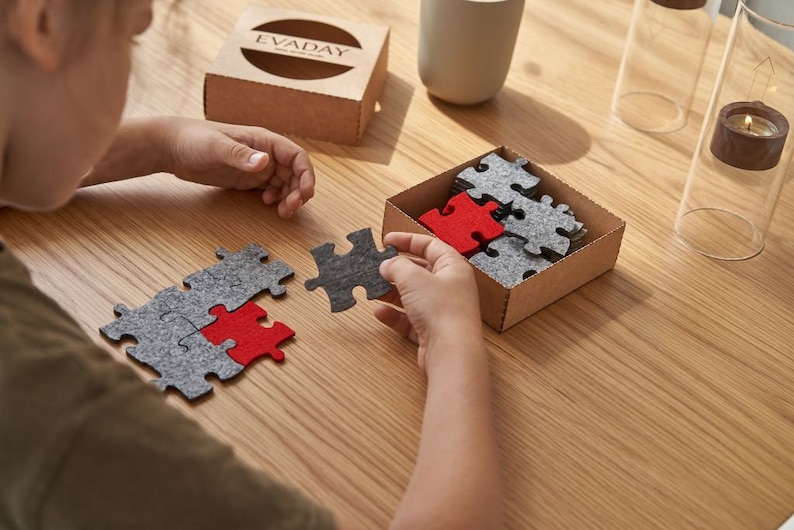 A child assembling grey and red felt puzzle pieces on a wooden table, with a box labeled "EVADAY" in the background.