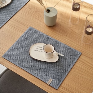 Può includere: Un tavolo di legno apparecchiato con due tovagliette grigie con un motivo geometrico. Ogni tovaglietta ha un piatto bianco, una tazza e un cucchiaio. Sul tavolo sono presenti anche candele decorative in portacandele di vetro e un vaso con steli secchi.