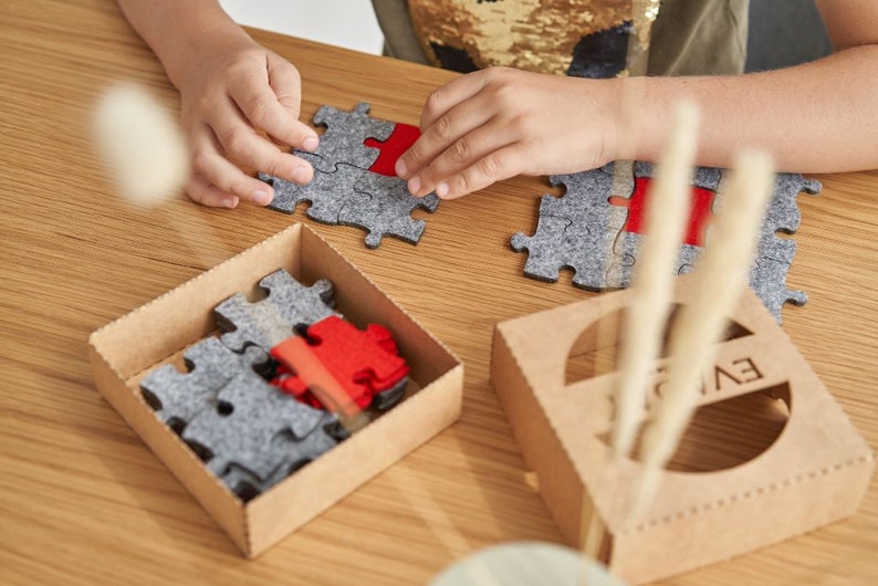 Overhead view of a child fitting grey and red felt puzzle pieces together on a wooden table.