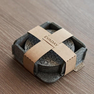 Puede incluir: Un juego de seis posavasos de fieltro gris con un patrón geométrico, empaquetados en una caja de fieltro gris con un envoltorio de papel marrón. El envoltorio tiene la palabra "EVADAY" impresa.