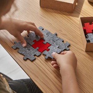 A child playing with a set of grey and red felt puzzle pieces on a wooden table, with a cozy setup including a mug and decorative items.