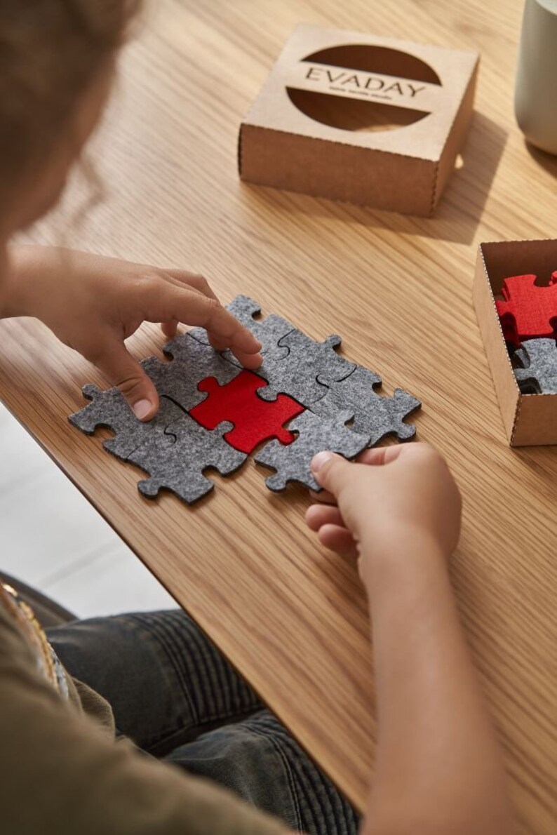 A child playing with a set of grey and red felt puzzle pieces on a wooden table, with a cozy setup including a mug and decorative items.