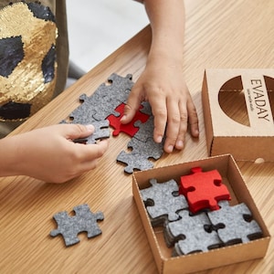 A child playing with grey and red felt puzzle pieces on a wooden table, with a mug and the "EVADAY" box nearby.