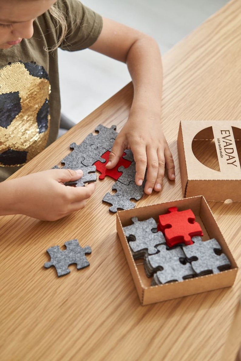 A child playing with grey and red felt puzzle pieces on a wooden table, with a mug and the "EVADAY" box nearby.