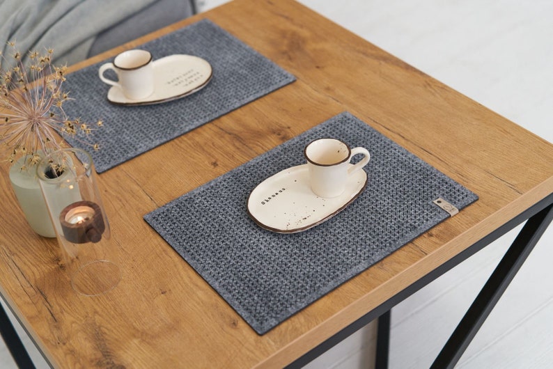 A stylish dining setup with a wooden table, grey textured placemats, white ceramic plates, and small coffee cups, adorned with simple decorative pieces.