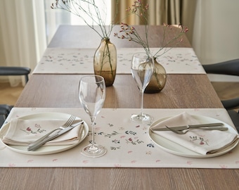 May include: Two place settings with white plates, napkins, and silverware on a light brown table runner with a floral pattern. Two clear glass vases with dried flowers are on the table.
