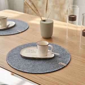 以下が含まれることがあります： 明るい木製のテーブルの上に、幾何学模様のグレーの丸いプレースマットが2つ置かれています。1つのプレースマットの上に、白いセラミック製のカップとソーサーが置かれています。テーブルの上には、火が灯ったキャンドルが入ったガラス製のキャンドルホルダーが2つあります。