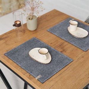 A neatly arranged wooden table with grey textured placemats, white ceramic plates, and small coffee cups, viewed from above.