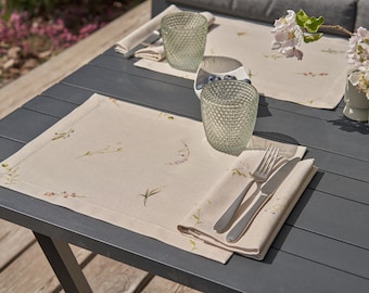 May include: Two place settings on a gray table with a floral pattern placemat, a white napkin, silverware, and a clear glass. The table is outdoors on a wooden deck.
