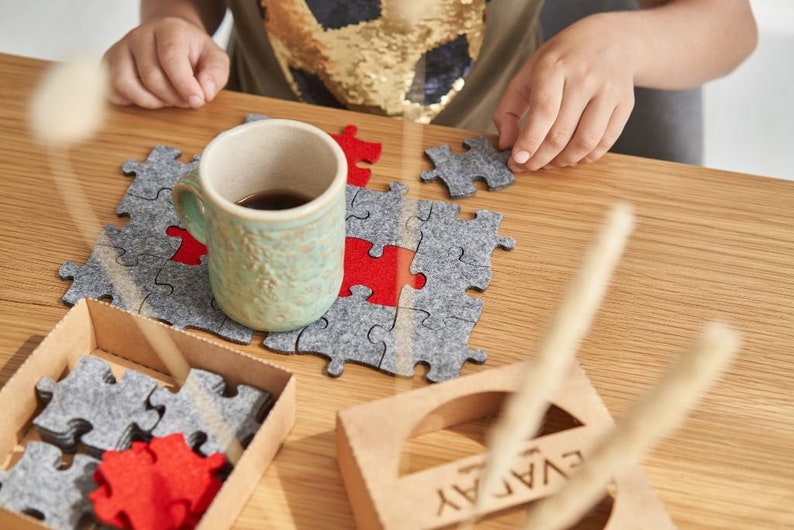 A child creating a pattern with grey and red felt puzzle pieces on a wooden table, accompanied by a mug and the "EVADAY" box.