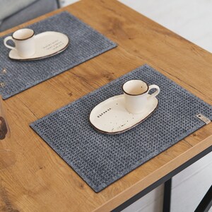 A wooden table set for two with grey textured placemats, white ceramic plates, and small coffee cups, accompanied by decorative items.