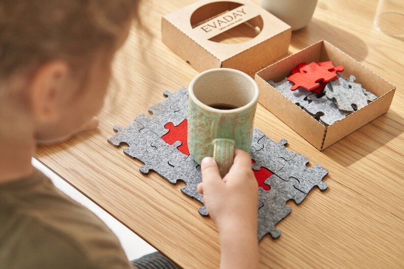 A child enjoying assembling grey and red felt puzzle pieces on a wooden table, with a mug in the background.