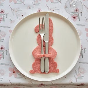 May include: A white plate with a pink felt bunny-shaped utensil holder, holding a fork and knife. The table setting is on a floral patterned tablecloth with pink flowers and green leaves. A wine glass is visible in the background.