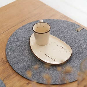 May include: A white ceramic cup and saucer with brown trim and speckles. The saucer has the word "Starry" written on it. The cup and saucer are on a grey round felt coaster.