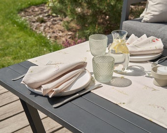 May include: A table setting with a white tablecloth with floral print, two glasses, a pitcher of water with lemon, a folded napkin, a plate, a fork, a knife, and a candle holder with a white candle.