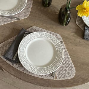Peut inclure: Vue aérienne d'une table ronde en bois dressée pour un repas. Des assiettes blanches aux bords décoratifs sont posées sur des sets de table à motifs. Des serviettes grises et des couverts sont disposés autour des assiettes. Un vase avec une fleur jaune est au centre.