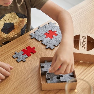 Close-up of a child arranging grey and red felt puzzle pieces on a wooden table, focusing on the puzzle pieces.