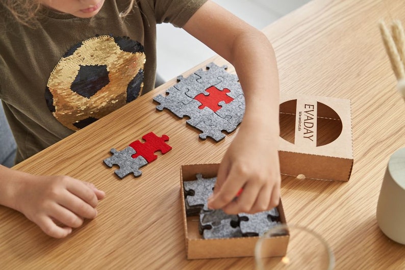 Close-up of a child arranging grey and red felt puzzle pieces on a wooden table, focusing on the puzzle pieces.